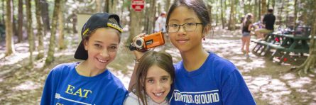 three campers smiling at camera while one of them holding a video camera