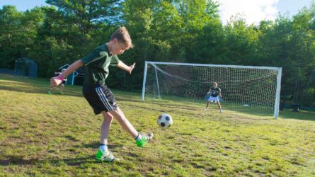 boy kicking a soccer ball to a goal while another camper defends
