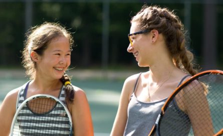 two young girls holding tennis rackets and smiling at each other